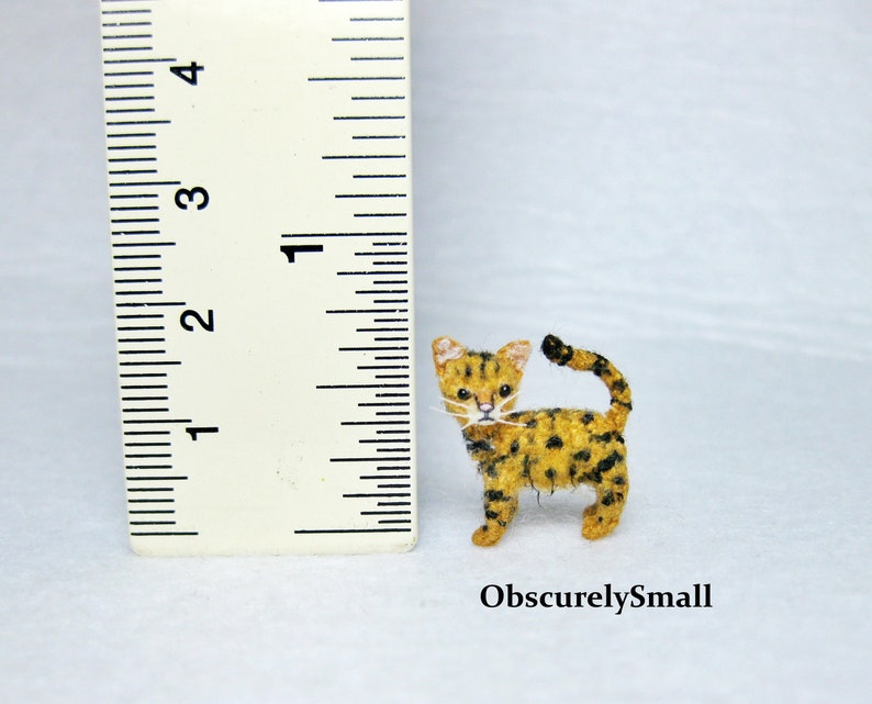 Pu&ograve; includere: Una piccola figurina di gatto in feltro fatta a mano con un motivo a macchie nere e gialle. Il gatto &egrave; in piedi su una superficie bianca accanto a un righello, misurando circa 2,5 cm di altezza.