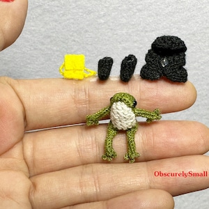 May include: A green crocheted frog with white accents, a yellow crocheted backpack, and two black crocheted bags. The items are arranged on a hand.