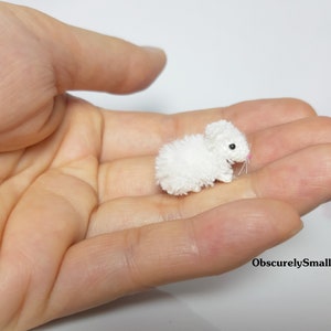 May include: A small, white crocheted bunny rabbit with pink nose and black eyes. The bunny is sitting in the palm of a hand.