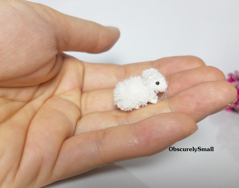 May include: A small, white crocheted bunny rabbit with pink nose and black eyes. The bunny is sitting in the palm of a hand.
