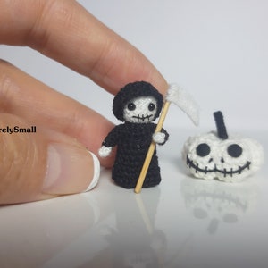 May include: A small crocheted Grim Reaper holding a scythe, standing next to a crocheted pumpkin with a smiling face.