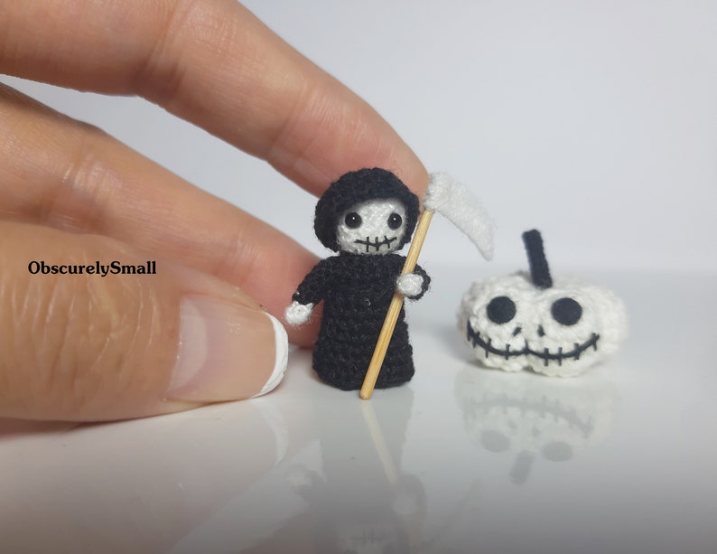 May include: A small crocheted Grim Reaper holding a scythe, standing next to a crocheted pumpkin with a smiling face.