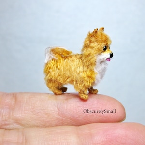 May include: A small, crocheted, orange and white Pomeranian dog figurine. The dog is standing on a finger and has a pink tongue sticking out.