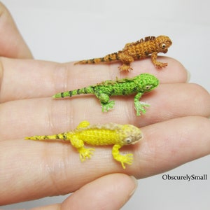 Op de afbeelding: Drie miniatuurgebreide hagedissen in verschillende kleuren: groen, geel en oranje. De hagedissen zijn geposeerd op een hand, waardoor hun ingewikkelde details worden getoond.
