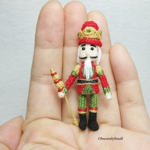 May include: A miniature crocheted nutcracker doll with a red and green uniform, a gold crown, and a white beard. The doll is holding a gold and red stick.