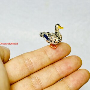 Op de afbeelding: Een klein, handgestikt eendje met een grijs en wit gepatroneerd lichaam, blauwe vleugels en een gele snavel. De eend staat op een vinger.