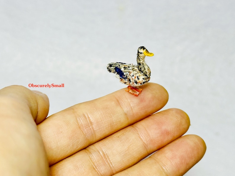 Op de afbeelding: Een klein, handgestikt eendje met een grijs en wit gepatroneerd lichaam, blauwe vleugels en een gele snavel. De eend staat op een vinger.