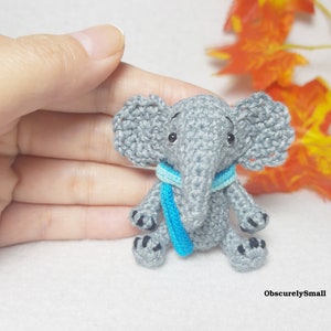 May include: A small gray crocheted elephant figurine featuring large textured ears, black bead eyes, and a bright blue scarf wrapped around its neck. The elephant has black stitched details on its feet and the text ObscurelySmall in the corner.