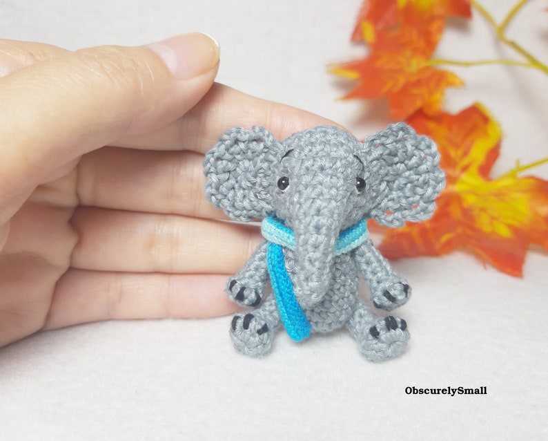 May include: A small gray crocheted elephant figurine featuring large textured ears, black bead eyes, and a bright blue scarf wrapped around its neck. The elephant has black stitched details on its feet and the text ObscurelySmall in the corner.