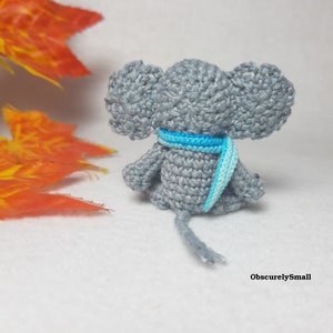 May include: A small, gray crocheted elephant figurine viewed from the back, featuring large ears and a bright blue and light blue striped scarf wrapped around its neck. The elephant has a thin, gray crocheted tail and the text ObscurelySmall.