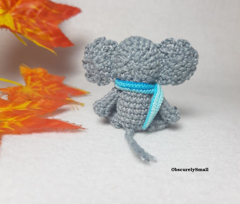 May include: A small, gray crocheted elephant figurine viewed from the back, featuring large ears and a bright blue and light blue striped scarf wrapped around its neck. The elephant has a thin, gray crocheted tail and the text ObscurelySmall.