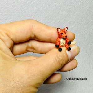 以下が含まれることがあります： オレンジと白の小さな編みぐるみキツネの置物。黒い目と鼻をしています。キツネは尻尾を後ろに巻いて座っています。置物は人の手に持たれています。