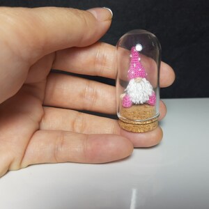 May include: A small gnome figurine with a pink and white hat sits inside a clear glass jar with a cork stopper. The gnome is wearing a white beard and pink trousers.