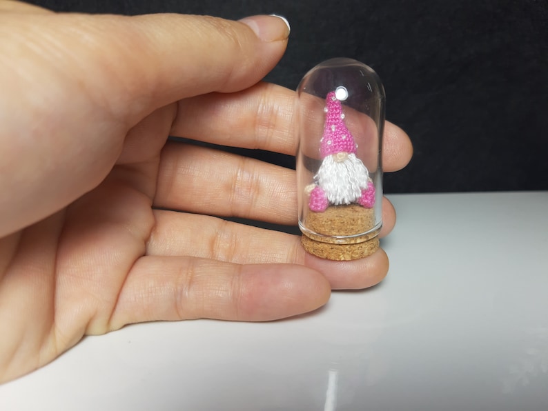 May include: A small gnome figurine with a pink and white hat sits inside a clear glass jar with a cork stopper. The gnome is wearing a white beard and pink trousers.