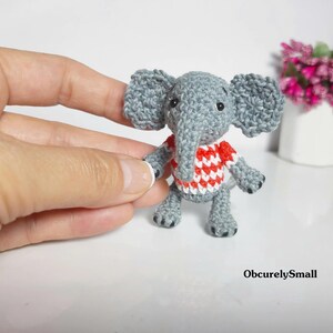 May include: A small, gray crocheted elephant figurine featuring large ears, a trunk, and a red and white checkered shirt. The elephant has black bead eyes and dark gray stitched details on its feet. The text ObcurelySmall is visible.