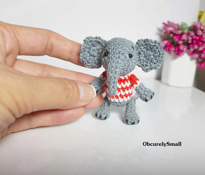 May include: A small, gray crocheted elephant figurine featuring large ears, a trunk, and a red and white checkered shirt. The elephant has black bead eyes and dark gray stitched details on its feet. The text ObcurelySmall is visible.