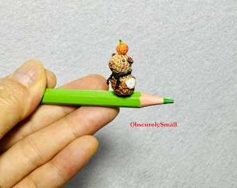 May include: A tiny crocheted animal wearing a scarf and holding a small orange fruit on top of its head. The animal is sitting on a green pencil.