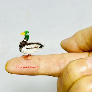Op de afbeelding: Een klein, handgemaakt, gehaakt wilde eendje met een groene kop, bruine en witte veren en oranje poten. De eend zit op een vinger.