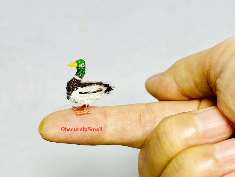 Op de afbeelding: Een klein, handgemaakt, gehaakt wilde eendje met een groene kop, bruine en witte veren en oranje poten. De eend zit op een vinger.