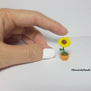 May include: A miniature crocheted sunflower in a small orange pot. The sunflower has bright yellow petals and a brown center. The green stem and leaves add contrast. The pot is a woven design. The text "ObscurelySmall" is in the bottom right corner.