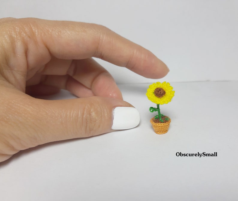 May include: A miniature crocheted sunflower in a small orange pot. The sunflower has bright yellow petals and a brown center. The green stem and leaves add contrast. The pot is a woven design. The text "ObscurelySmall" is in the bottom right corner.