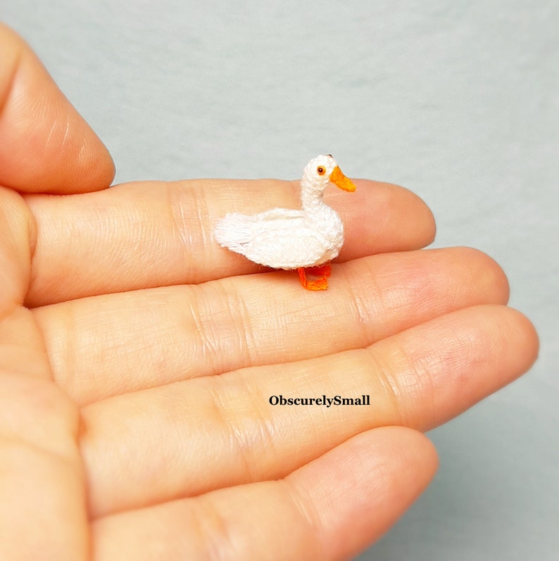 May include: A small, white crocheted duck with orange feet and a black eye. The duck is sitting on a person's hand.