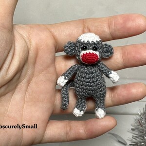Puede incluir: Un peque&ntilde;o mono de crochet gris con detalles blancos y una nariz roja. El mono est&aacute; sentado en una mano.