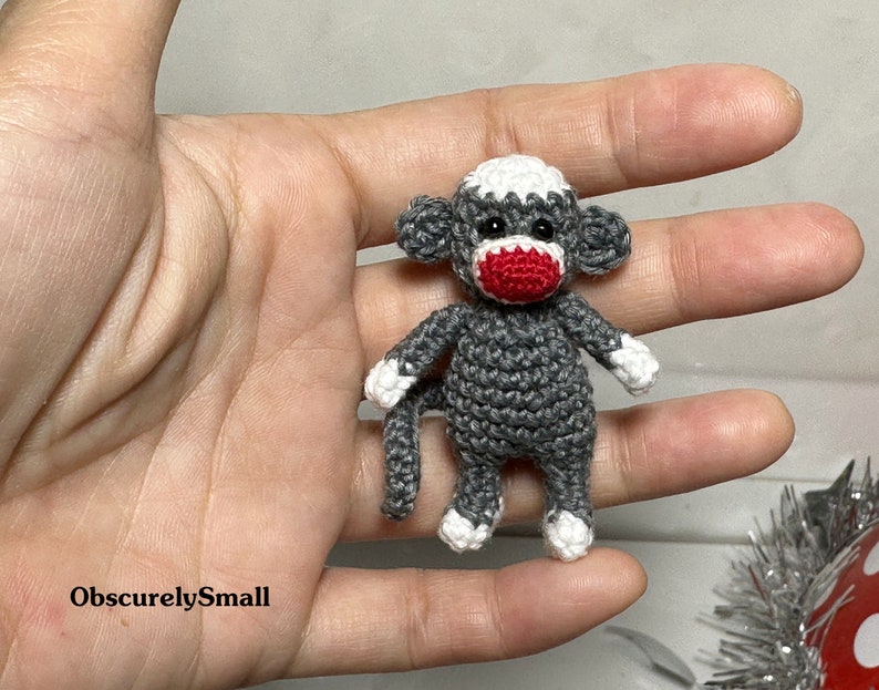 Puede incluir: Un peque&ntilde;o mono de crochet gris con detalles blancos y una nariz roja. El mono est&aacute; sentado en una mano.