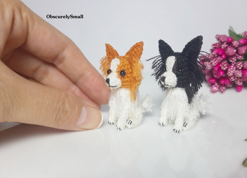May include: Two small crocheted Papillon dogs, one with orange and white fur and the other with black and white fur. Both dogs are sitting with their heads turned to the side.