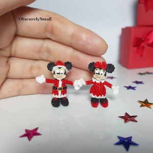 Puede incluir: Figuras de ganchillo de Mickey Mouse y Minnie Mouse vestidas con trajes de Papá Noel. Mickey lleva un traje de Papá Noel rojo y blanco con un cinturón negro y zapatos negros. Minnie lleva un vestido rojo y blanco con un lazo blanco y zapatos negros. Las figuras están de pie sobre una superficie blanca.