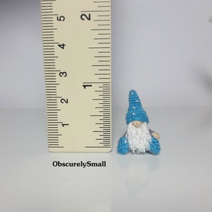 May include: A small crocheted gnome wearing a blue hat with white polka dots and a white beard. The gnome is sitting on a white surface with a ruler in the background.