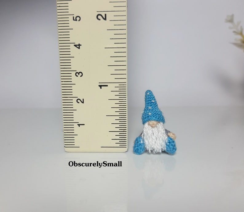 May include: A small crocheted gnome wearing a blue hat with white polka dots and a white beard. The gnome is sitting on a white surface with a ruler in the background.