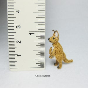 May include: A small crocheted kangaroo toy, standing about 1 inch tall, next to a ruler. The kangaroo is brown with white accents and is holding a smaller crocheted kangaroo in its pouch.
