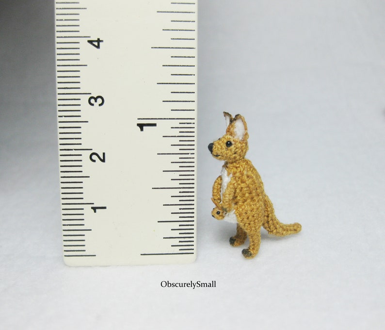 May include: A small crocheted kangaroo toy, standing about 1 inch tall, next to a ruler. The kangaroo is brown with white accents and is holding a smaller crocheted kangaroo in its pouch.