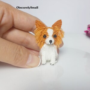 May include: A small, crocheted, white and orange papillon dog figurine. The dog is sitting with its ears perked up and its tail curled.