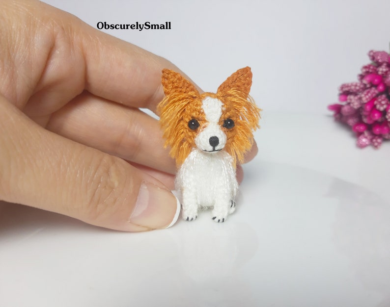 May include: A small, crocheted, white and orange papillon dog figurine. The dog is sitting with its ears perked up and its tail curled.