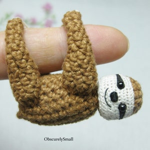 May include: A small crocheted sloth hanging from a finger. The sloth is brown with a white face and black eyes. The sloth is about the size of a finger.