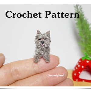 Könnte beinhalten: Häkelmuster für einen kleinen, grauen, gehäkelten Hund, der auf einem Finger sitzt. Der Hund hat eine rosa Zunge herausgestreckt. Der Text "Crochet Pattern" befindet sich oben im Bild. Der Text "ObscurelySmall" befindet sich unten im Bild.