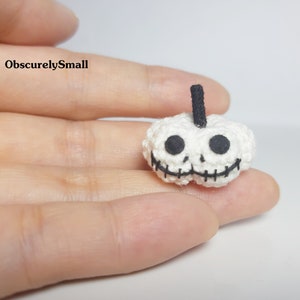 May include: A small, white crocheted pumpkin with a black stem and a skull face. The eyes and mouth are black, stitched details. The text "ObscurelySmall" is in the upper left corner.