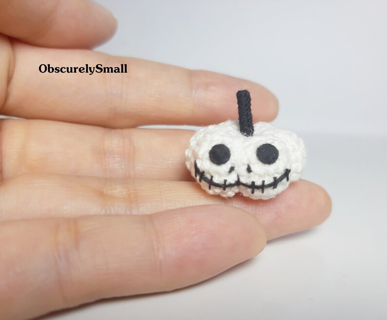 May include: A small, white crocheted pumpkin with a black stem and a skull face. The eyes and mouth are black, stitched details. The text "ObscurelySmall" is in the upper left corner.