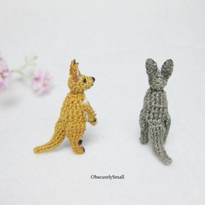 May include: Two crocheted kangaroo figurines, one is a light brown color and the other is a gray color. Both are standing upright with their tails curled behind them.