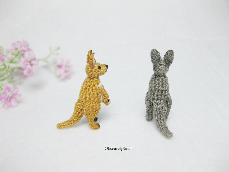 May include: Two crocheted kangaroo figurines, one is a light brown color and the other is a gray color. Both are standing upright with their tails curled behind them.
