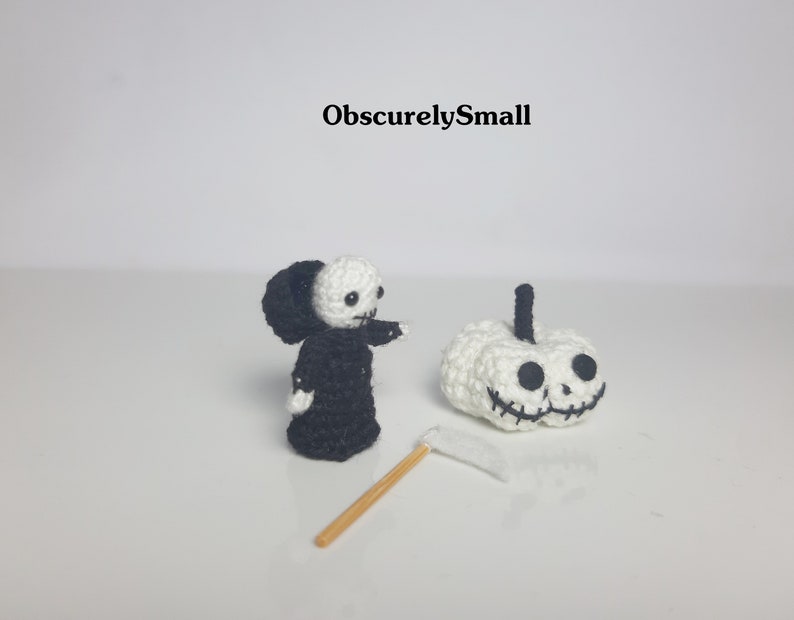 May include: A crocheted Grim Reaper figure in a black robe with a white face and a white pumpkin with a black face and a stitched smile. The Grim Reaper is holding a small white scythe.
