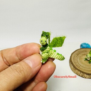 May include: A small green crocheted dragon with white accents. The dragon has a wing and is standing on a hand.