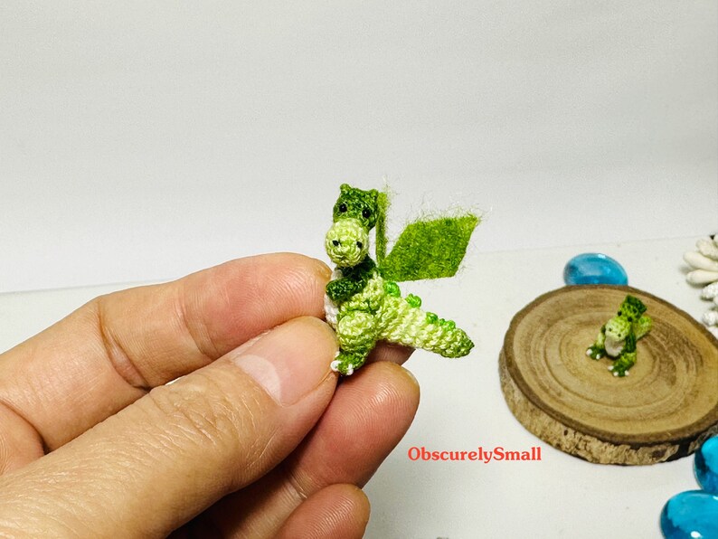 May include: A small green crocheted dragon with white accents. The dragon has a wing and is standing on a hand.