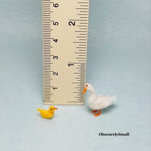 May include: Two crocheted miniature ducks, one white and one yellow, are shown next to a ruler. The white duck is about 1 inch tall and the yellow duck is about 1/2 inch tall. The ducks are made with yarn and have small orange beaks and feet.