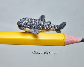 Puede incluir: Una pequeña figura de tiburón ballena de crochet está posada sobre un lápiz amarillo. El tiburón es gris con manchas blancas y tiene la boca ligeramente abierta.