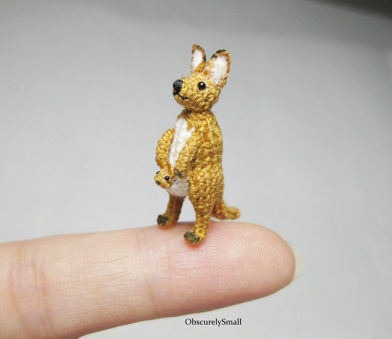 May include: A small crocheted kangaroo with a joey in its pouch. The kangaroo is brown and white, and the joey is white. The kangaroo is standing on a finger.