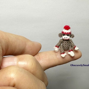 May include: A tiny crocheted monkey wearing a red and white hat. The monkey is sitting on a finger.
