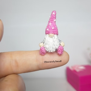 May include: A tiny crocheted gnome with a pink hat with white polka dots, a white beard, and pink shoes. The gnome is sitting on a finger.